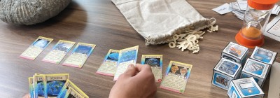 Photographie montrant une partie de jeu de cartes éducatif. Plusieurs cartes disposées en rangée et un sablier orange en haut à droite. Main humaine en train de manipuler une carte, plusieurs paquets de cartes empilés à droite, et un sac en tissu beige contenant des jetons en forme de lettres.